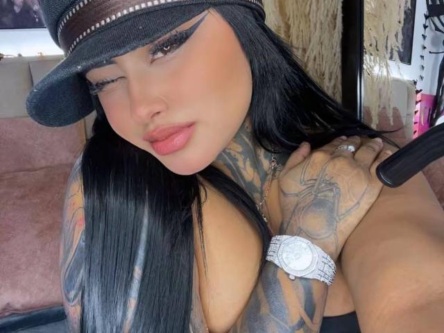 LairaHearvy with long black hair, wearing a hat, winking, showing tattoos, big tits, and a watch, with her hand on her shoulder, and pink lipstick.