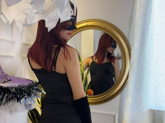 Sahara69 with red hair, black mask, black dress, and black gloves, reflected in a gold mirror. She's looking at herself, showing her tits and ass in the reflection.