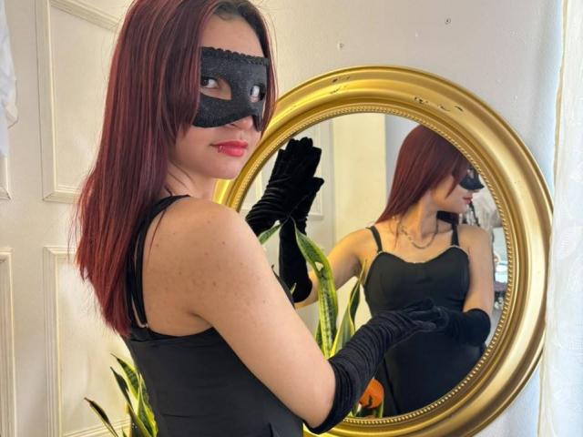 Sahara69 with red hair, black mask, black dress, and black gloves, reflected in a gold mirror, showing her tits and ass in the reflection.