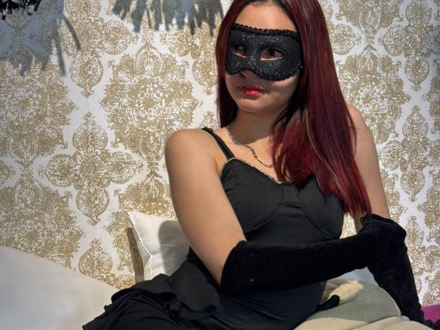Sahara69 with red hair, black mask, and black dress, wearing long black gloves, sitting on a bed with white pillows, smiling slightly, showing red lipstick.