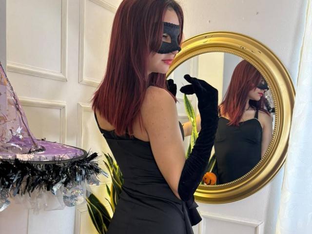 Sahara69 with red hair, black mask, black dress, and black gloves, standing in front of a gold mirror, showing her ass in the reflection. Her tits are covered.