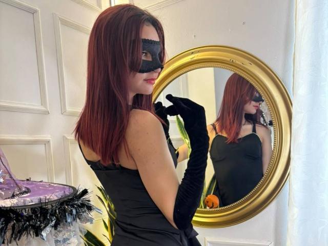 Sahara69 with red hair, black mask, black dress, and black gloves, standing in front of a gold mirror, showing her tits in the reflection, looking sexy.