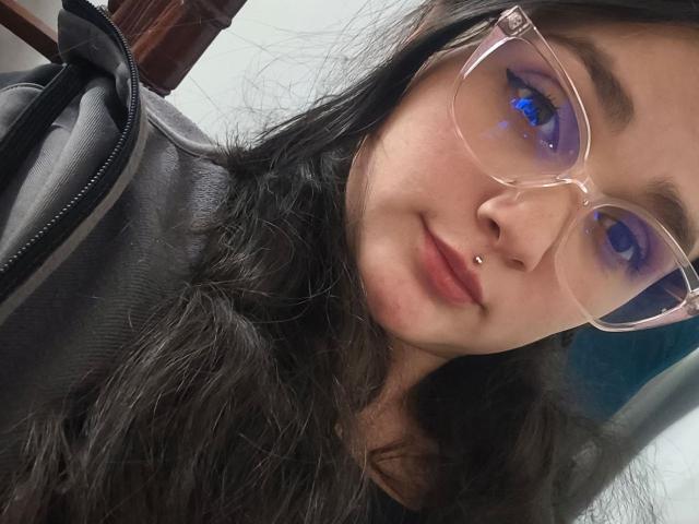 NataliaRivas, avec du fard à paupières violet, des lunettes transparentes, des cheveux noirs ondulés, un piercing au nez, et une veste en cuir noir, affichant un léger sourire, regardant directement l'appareil photo..