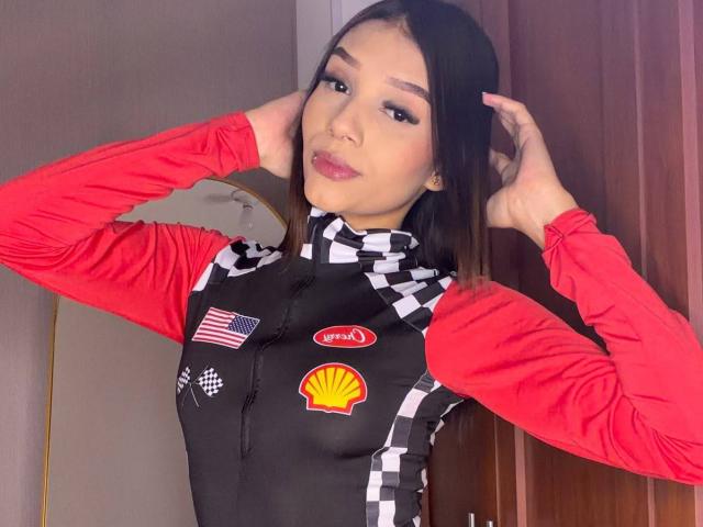 SaraMillerOne with long brown hair, wearing a red and black checkered racing vest, hands in hair, looking at camera, American flag and Shell logos on vest.