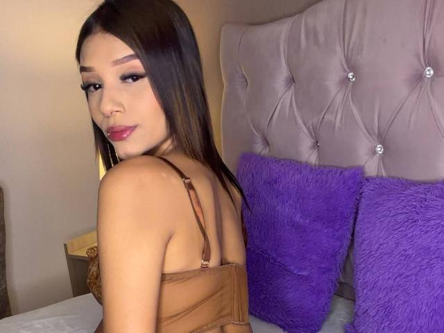 SaraMillerOne with brown bra, purple pillows, and tufted headboard, looking over shoulder, showing her back and small tits, brown hair, pink lipstick, and subtle makeup.