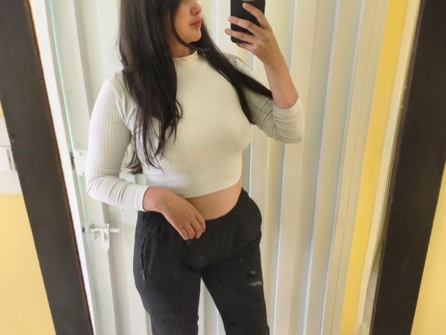 NataliaRivas mit langen, dunklen Haaren, einem weißen, gerippten Crop-Top, schwarzen Hosen, die ein Handy vor einem Spiegel hält. Sichtbare Brüste, weicher Bauch, leicht kurvig, stehend, gelbe und weiße Wand..