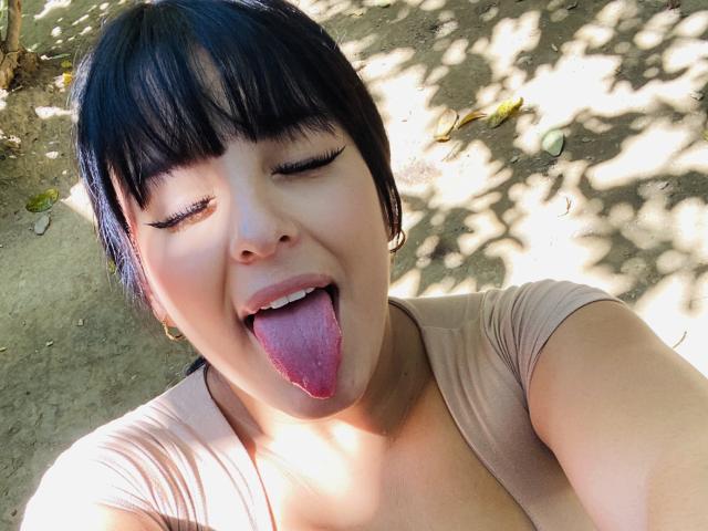 TalianaSquirt has short black hair, closed eyes, and her tongue out, wearing a beige top, with sunlight casting shadows on her face. Her pink tongue is prominently visible.