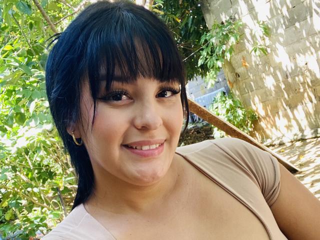 TalianaSquirt with black hair, bangs, and brown eyes, smiling with full lips, wearing a beige top, showing a chubby face and small, round tits, in a sunny outdoor setting.