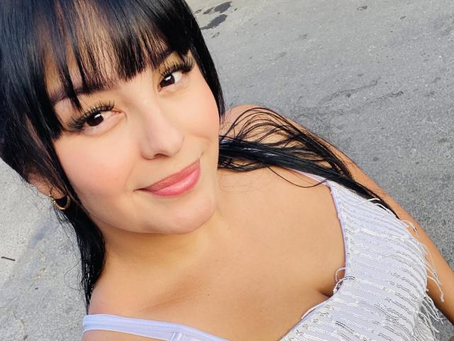 TalianaSquirt with long black hair, bangs, and eyelashes, smiling, wearing a fringed white top, showing her tits, standing on a grey pavement, wearing small gold earrings.