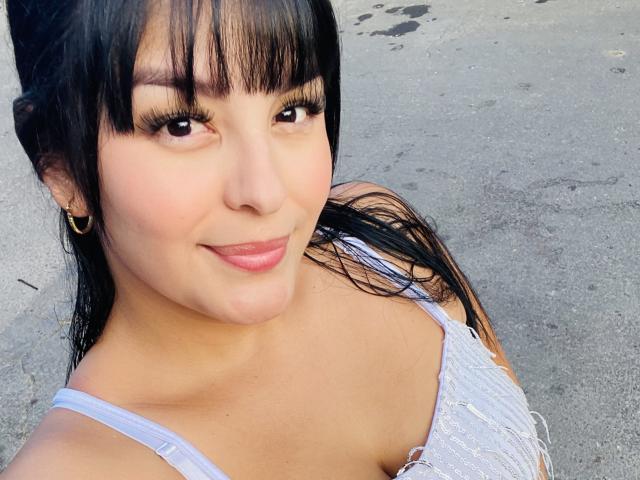 TalianaSquirt with black bangs, long eyelashes, and a subtle smile, wearing a grey, fringed top, showing her shoulders and small, cute tits. She has a tan, smooth face.