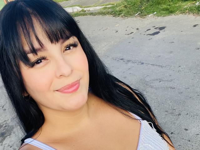 TalianaSquirt with long black hair, bangs, and a cute smile, showing off her pouty pink lips, wearing a white top, with her ass not visible in the image.