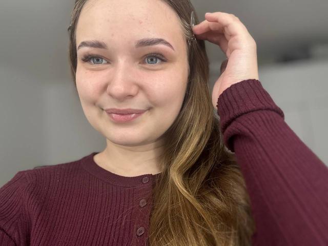 MagikPeachh with light skin, blue eyes, and long brown hair in a maroon rib-knit sweater, smiling slightly, hand in hair, showing natural, unshaved eyebrows and subtle pink lips.