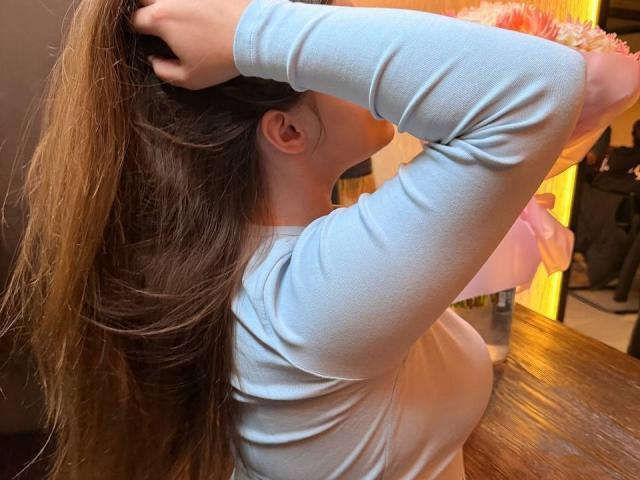 MagikPeachh with long brown hair, wearing a light blue long-sleeve shirt, hands in hair, showing her tits, standing by a wooden table in a brightly lit room.