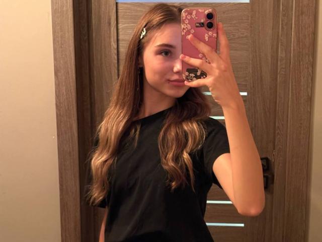 YanaPavlosky with long, wavy brown hair, wearing a black shirt, taking a mirror selfie with a pink phone, holding it in her right hand, showing a slight smile.