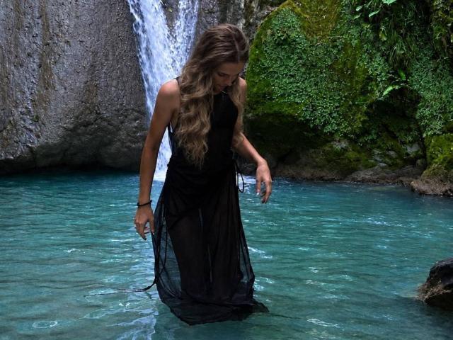 YanaPavlosky stands in clear blue water, wearing a black, sheer dress that reveals her tits and pussy, with long, wavy brown hair, waterfall in background.