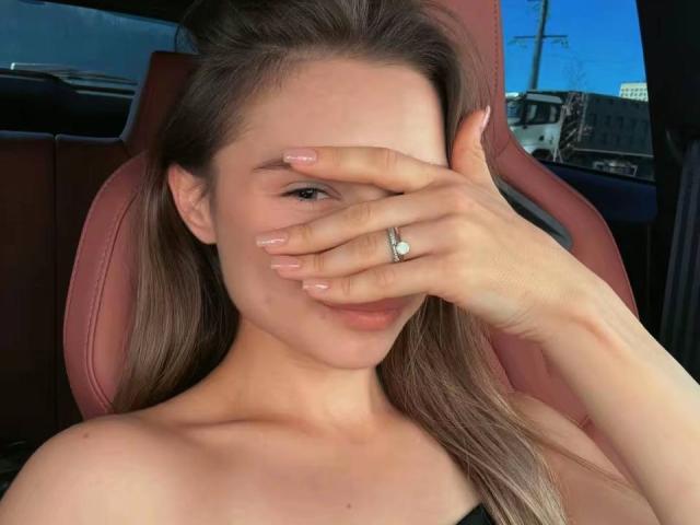YanaPavlosky with long brown hair, covering her eyes with her hand, showing a ring on her finger, sitting in a car with pink leather seats. Her tits are visible.