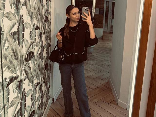 YanaPavlosky, mirror selfie, black sweater with white lines, gray pants, black bag, long brown hair, wooden floor, leaf pattern wall, holding phone, looking at mirror, confident.