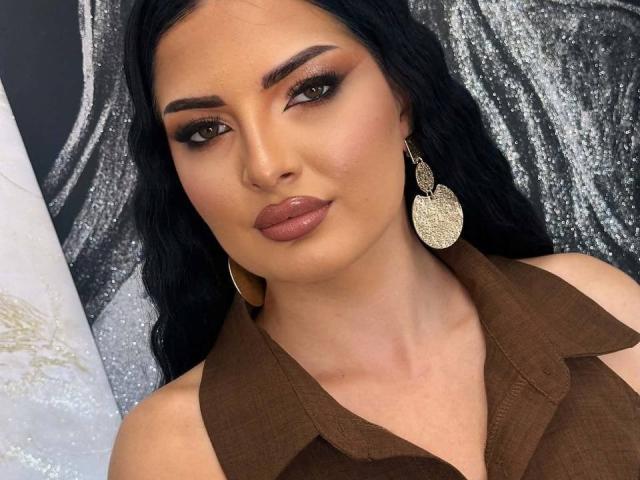 VelvetKaia with dark hair, brown shirt, gold earrings, and glossy pink lips. She's got thick eyebrows, subtle makeup, and a slight smile, looking straight at the camera.
