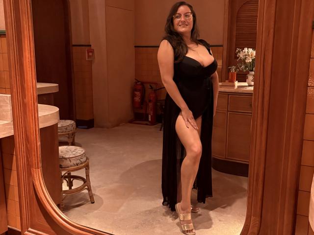 BadAngel with big boobs, dark hair, wearing a black dress with a high slit, standing in a wooden room, smiling, wearing clear heels, reflected in a mirror.