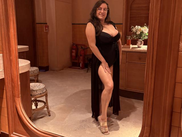 BadAngel with big boobs in a black dress, high leg slit, showing her ass, standing in front of a mirror, wearing gold heels, smiling, dark hair, glasses.
