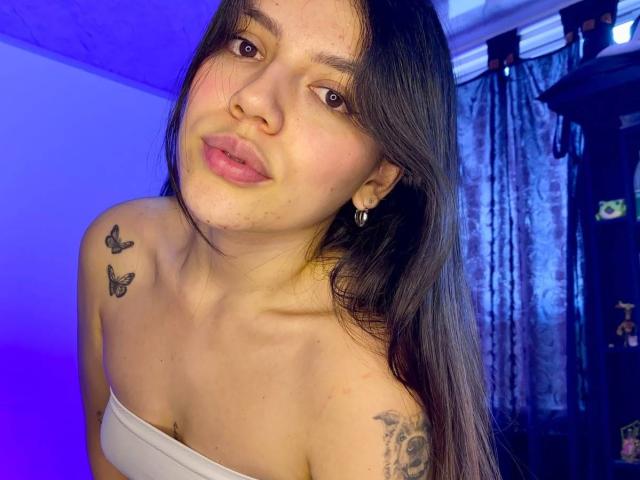 SophiaMarx with long black hair, pink lips, and small nose, wearing a white strapless top, showing butterfly tattoos on her shoulders, and a skull tattoo on her right arm.