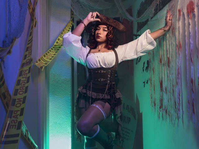 KanelaMiller, with curly brown hair, wearing a brown hat, white blouse, black corset, and frilled skirt, poses against a bloody wall, arms raised, in a dimly lit, green-blue room.