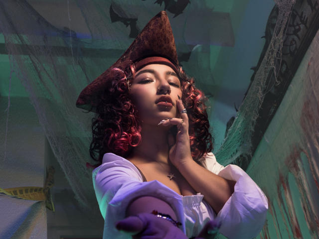 KanelaMiller with curly red hair, wearing a pirate hat, white shirt, and purple glove, touches her face, showing her tits and a necklace, with a confident, sultry expression.