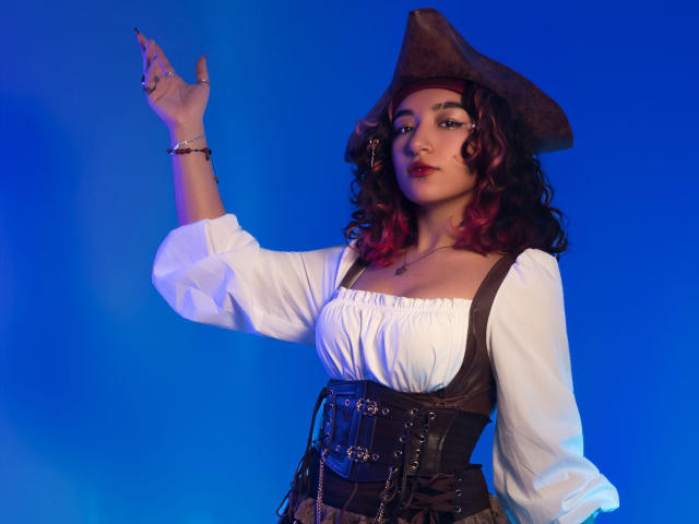 KanelaMiller with curly red hair, wearing a brown pirate hat, white blouse, brown corset, raising her right hand, blue background. Boobs visible under blouse, looking confident.