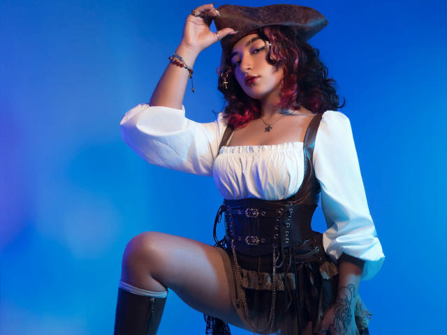 KanelaMiller with curly red-black hair, pirate hat, white puffy blouse, black corset, brown skirt, and knee-high boots, touching hat, showing tattoos, blue background, sexy pirate look.