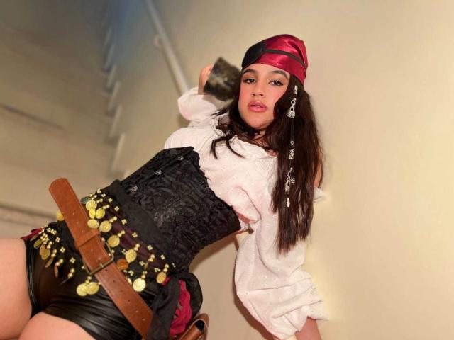 IrisBlair, dressed as a pirate, wears a red and black headscarf, white shirt, black corset, and leather shorts with gold coins and a brown belt, posing confidently.