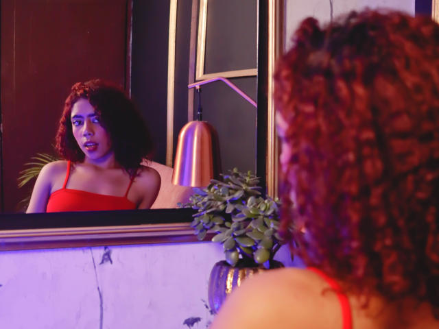Valerie Martin, avec ses cheveux roux bouclés, portant un débardeur rouge, regardant son reflet dans un miroir. Ses seins sont visibles, et une plante en pot se trouve au premier plan..