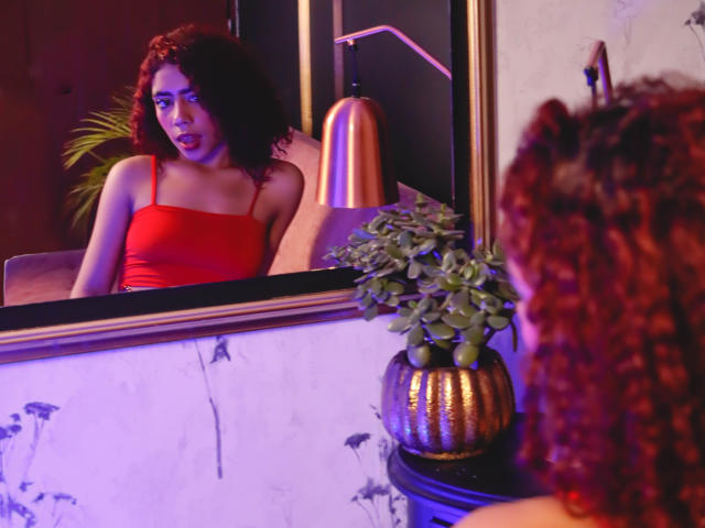 ValerieMartin with curly red hair, wearing a red tank top, looking at herself in a mirror. Her reflection shows her pouty lips and small tits. A plant is in front.