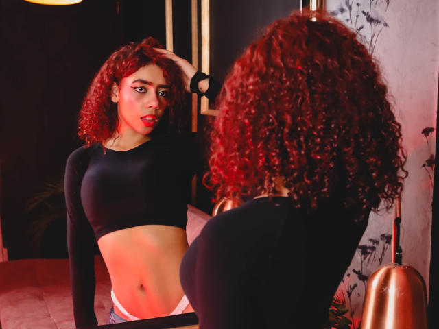 ValerieMartin with curly red hair, black crop top, showing her tits, wearing pink panties, standing in front of a mirror, hand in hair, red lighting, looking at reflection.