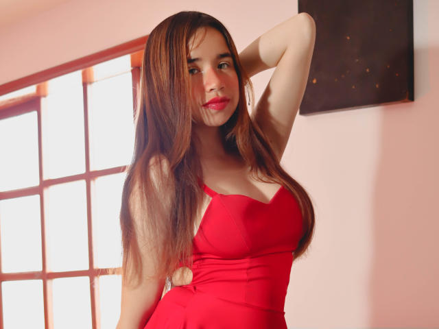 ValeriMonroe with long brown hair, red dress, and red lipstick, posing with arm behind head, showing her tits and small ass, in a well-lit room.