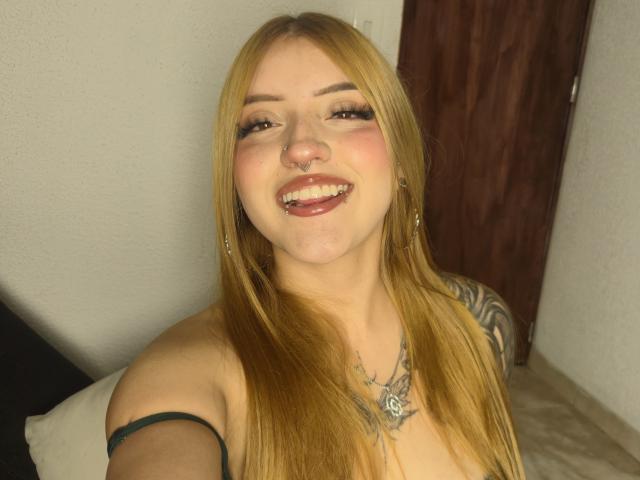 MailynnSkye with blonde hair, nose ring, tongue out, earrings, tattoos, and green strap, smiling at the camera, showing her tits and a nose piercing.