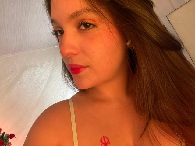 KatarinaNovak with long brown hair, red lipstick, small earrings, and a red tattoo on her chest, wearing a white spaghetti strap top, looking at the camera.