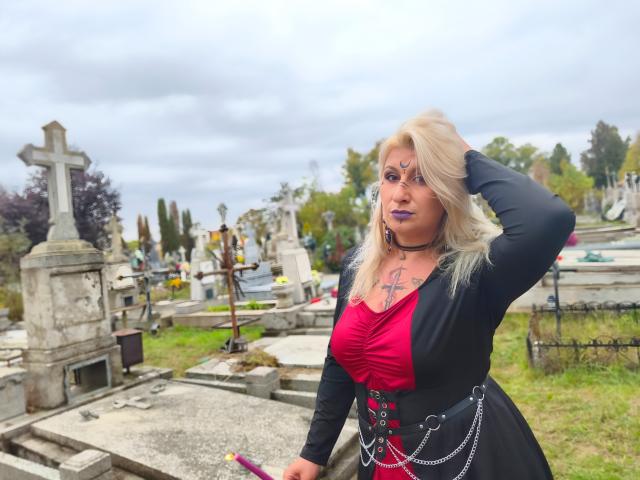 LilithGreenn has blonde hair, purple lipstick, and a black dress with a red bust, black corset, and chains. She's standing in a cemetery with her hand in her hair.