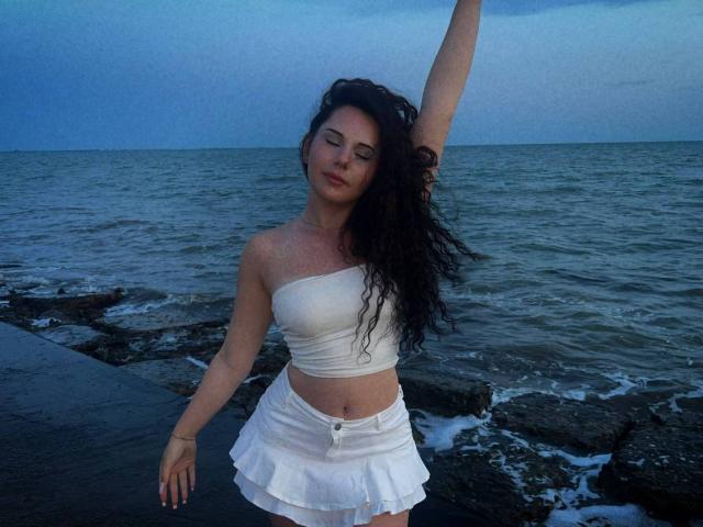 Laquita Zamborsky mit langem, lockigem, schwarzem Haar, einem weißen Top und einem weißen, gerüschten Rock, steht an einer felsigen Küste, ein Arm erhoben, die Augen geschlossen, ein leichtes Lächeln, im Hintergrund das Meer..