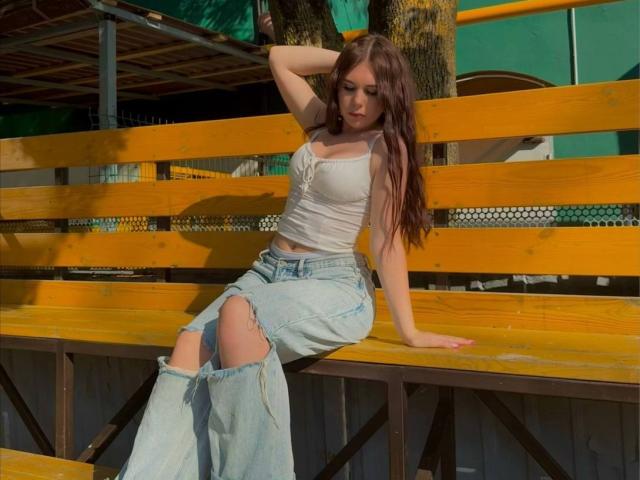 LaquitaZamborsky, with long brown hair, white tank top showing her tits, and ripped blue jeans, sits on a yellow wooden bench, arm behind her head, looking down.