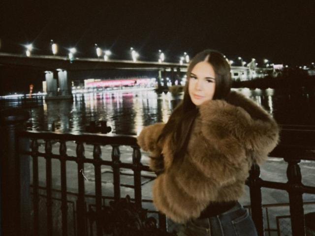 Laquita Zamborsky mit langen, braunen Haaren, die ein flauschiges, braunes Felljackett trägt, steht nachts an einem schwarzen Geländer, im Wasser spiegeln sich Lichter, im Hintergrund ist eine Brücke zu sehen..