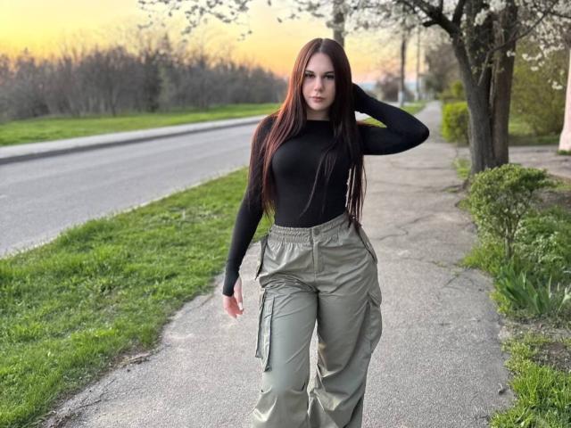 Laquita Zamborsky mit langen braunen Haaren, einem schwarzen Langarmshirt und grünen Cargohosen, steht auf einem Bürgersteig, die Hand im Haar, im Hintergrund ein Sonnenuntergang, links Gras und eine Straße..