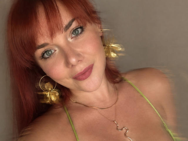 Dinastiia with red hair, green eyes, wearing gold earrings, hoop earrings, gold necklace, and green top, smiling with pink lipstick, showing a slight blur effect on her face.