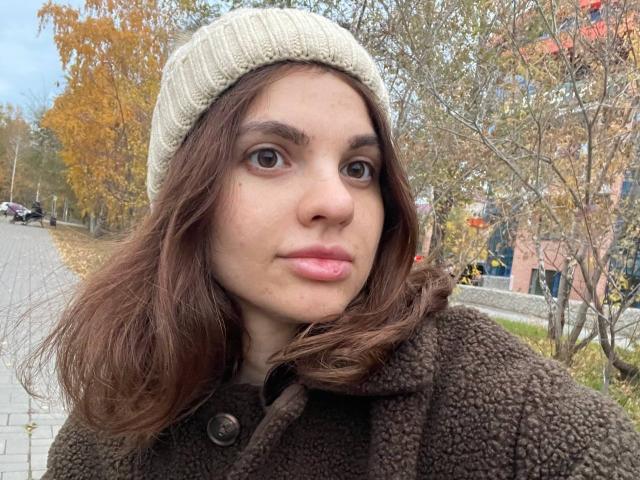 CicelyDence with wavy brown hair, wearing a beige knit hat and brown fuzzy coat, has puffy pink lips, brown eyes, and a slight nose bump, standing outdoors.