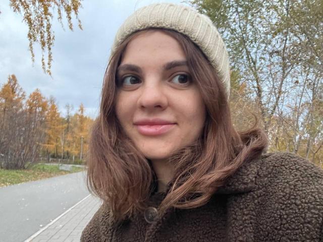 CicelyDence with wavy brown hair, wearing a beige knit cap and brown fuzzy coat, smiling slightly, with pink lips, in an autumn outdoor setting.