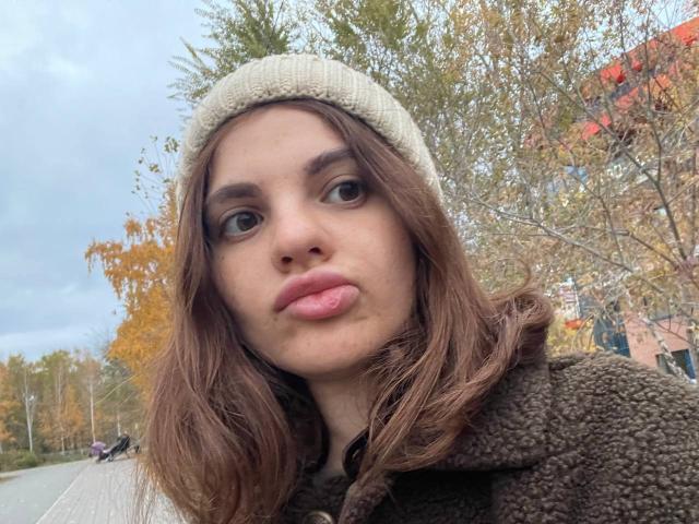 CicelyDence has brown hair, a beige knit beanie, and a fluffy brown coat. She's sticking her tongue out, with her nose and full lips visible. Autumn leaves in background.