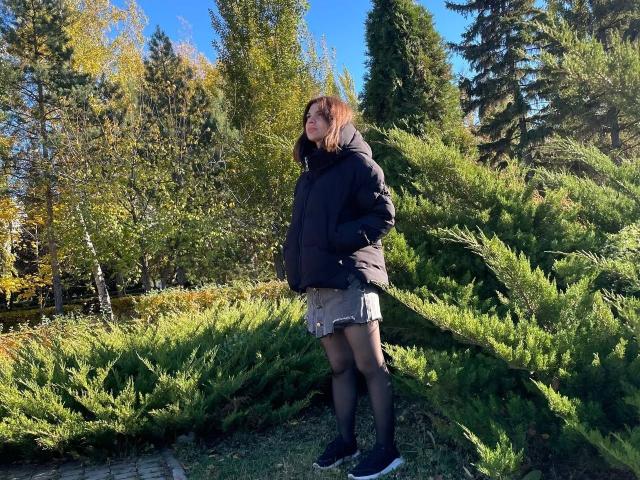 CicelyDence stands in a forest, wearing a black jacket, brown skirt, black tights, and black sneakers, with her hands in her jacket pockets, surrounded by evergreen trees.