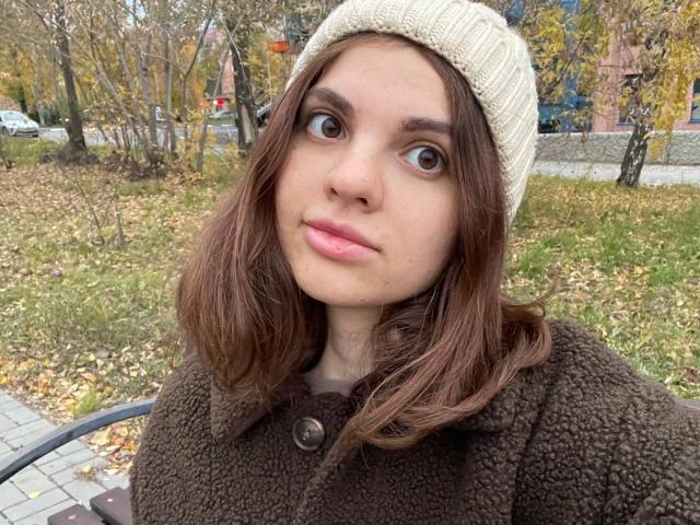 CicelyDence with wide eyes, pink lips, brown wavy hair, beige knit hat, brown fuzzy coat, buttoned up, standing in a grassy autumn area, looking directly at the camera.