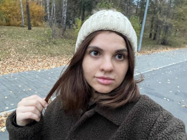 CicelyDence has brown eyes, a light skin tone, and wears a beige knitted beanie with a brown fuzzy coat, holding a lock of brown hair, looking at the camera.