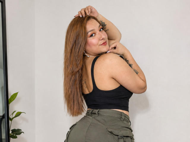 DuboisCarla with long brown hair, black tank top, and green pants, looking back, hand on head, showing tattoos, smiling with pink lips, butt prominent, white wall background.