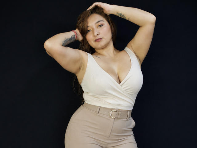 DuboisCarla with big tits, tan skin, brown hair, white tank top, beige pants, arms raised, tattoo on left arm, black background, confident expression.